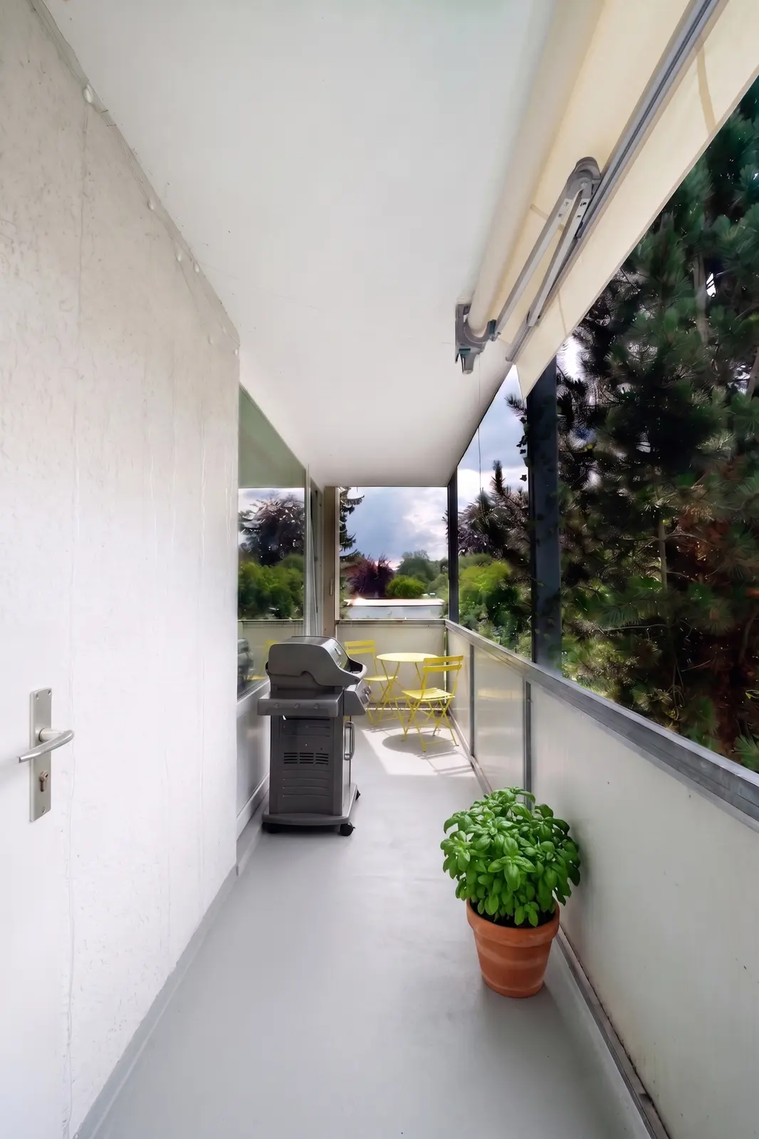 Ein schmaler Balkon mit einem Grill, einer Basilikumpflanze im Topf und einem kleinen gelben Tisch mit zwei Stühlen, von dem aus man durch ein Glasgeländer auf die grüne Umgebung blickt.