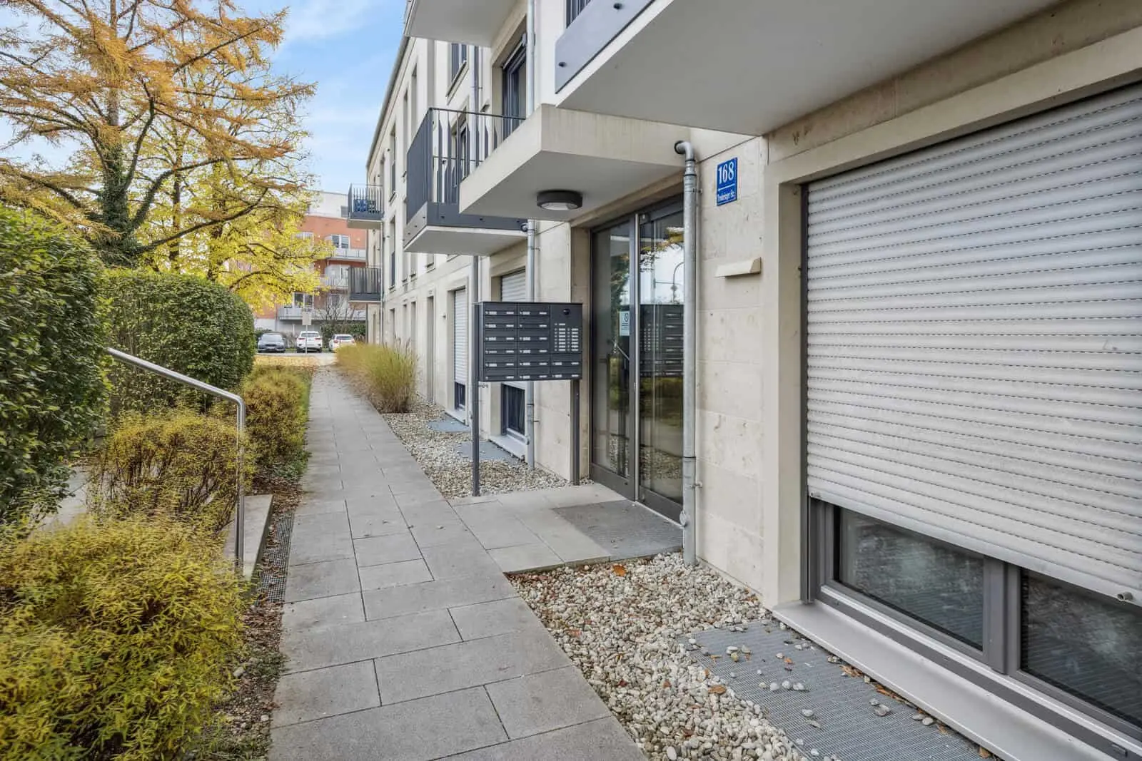 Moderner Wohnungseingang mit mehreren Briefkästen, Glastür und geschlossenen Fensterläden, umgeben von Sträuchern und einem gepflasterten Gehweg.