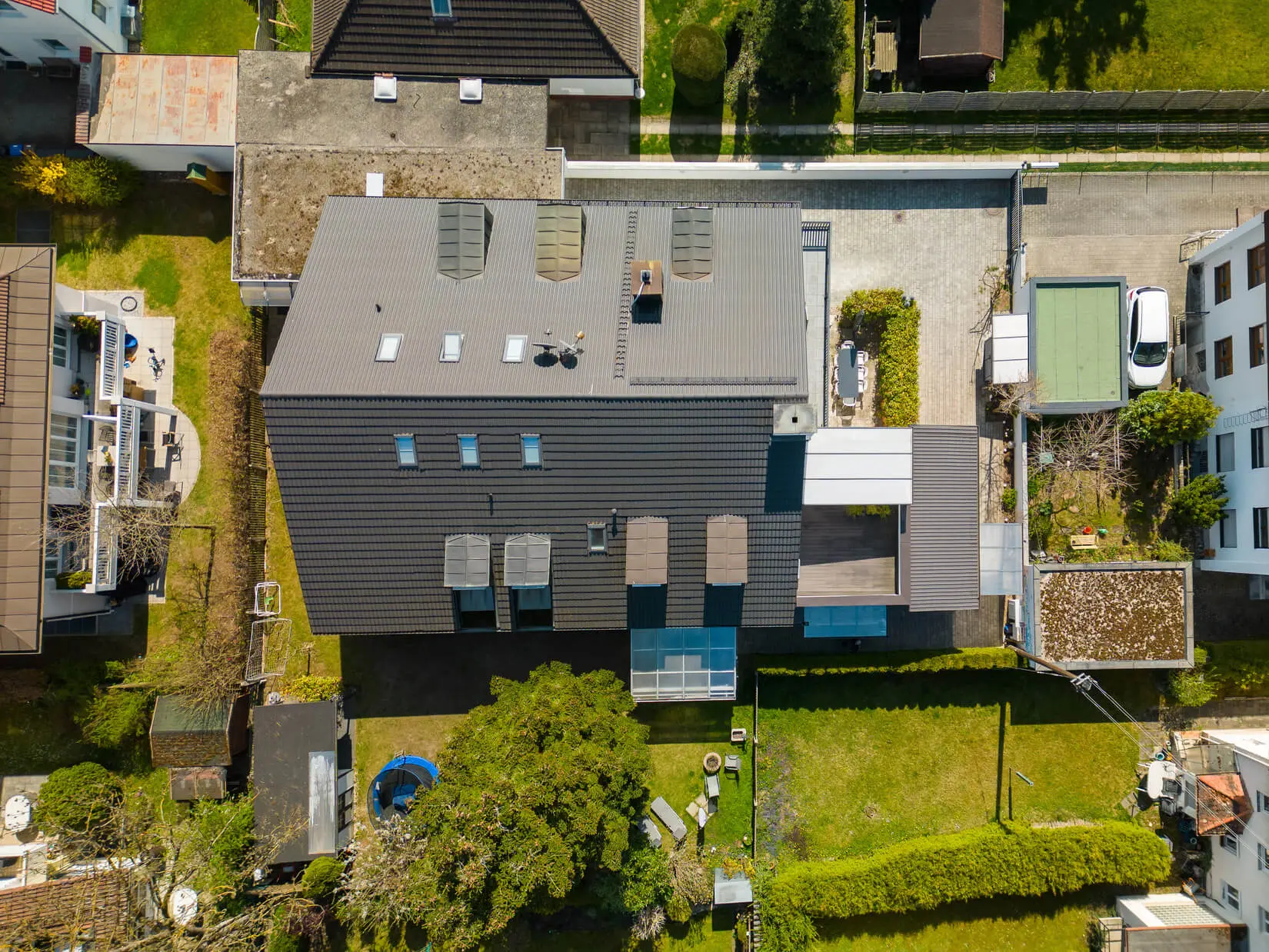 Luftaufnahme eines modernen, rechteckigen Hauses mit dunklem Dach, Oberlichtern und Außenbereichen, umgeben von einem grünen Rasen und Nachbargebäuden.