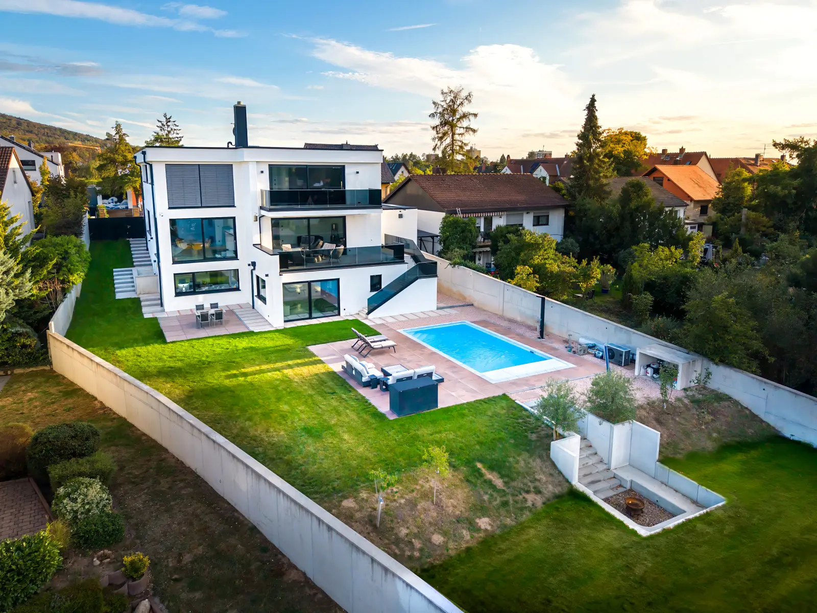 Modernes, weißes, mehrstöckiges Haus mit einem Swimmingpool im Hinterhof, Terrassenmöbeln und einem von einer Umfassungsmauer umgebenen Rasen.
