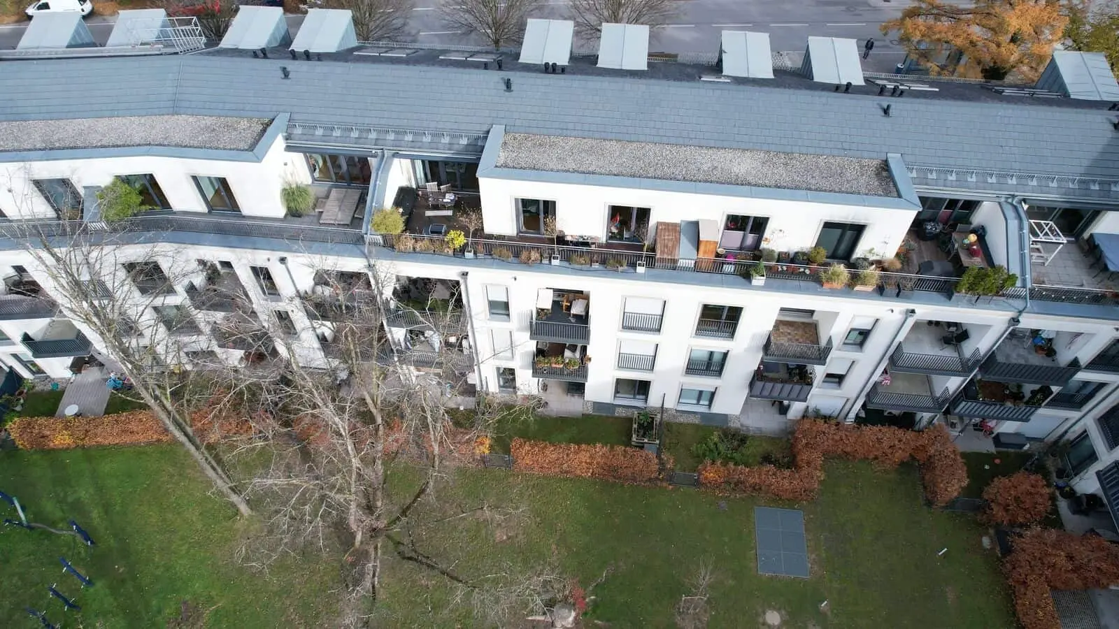 Luftaufnahme eines modernen Mehrfamilienhauses mit Balkonen, teilweise mit Pflanzen und Möbeln, mit Blick auf einen grasbewachsenen Hof mit Bäumen und Sträuchern.