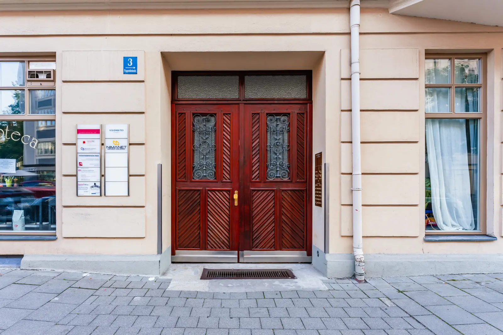 Rote Flügeltüren mit dekorativen schwarzen Metallpaneelen an einem beigen Gebäude, flankiert von Schildern und einem großen Fenster mit transparenten Vorhängen.