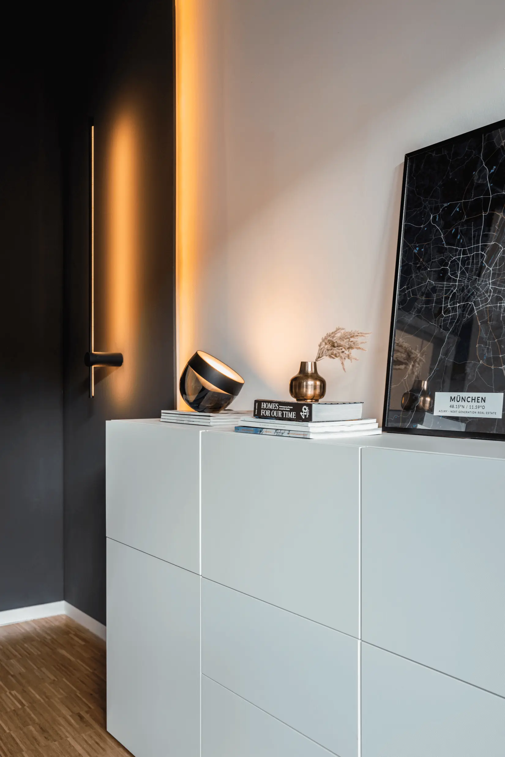 Minimalistisches Interieur mit weißen Schränken, modernen Deko-Artikeln und einer gerahmten Karte, die die Essenz von „Über uns“ verkörpert. Oben sitzt eine kleine Leuchte, während im Hintergrund eine Tür mit sanfter Beleuchtung teilweise sichtbar ist.